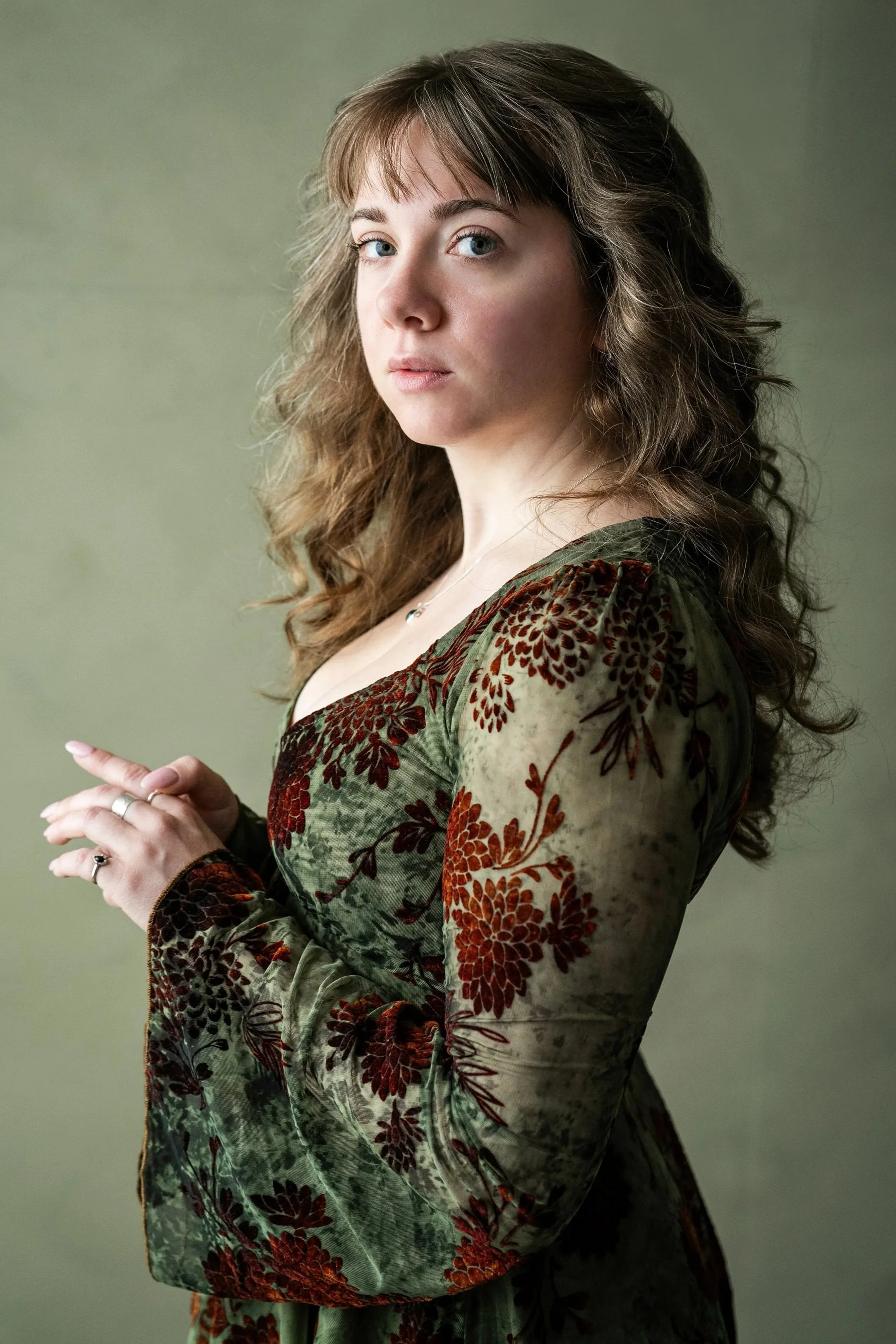 A woman with light skin and wavy brown hair, wearing a green dress with a red floral pattern, looks at the camera against a green background.