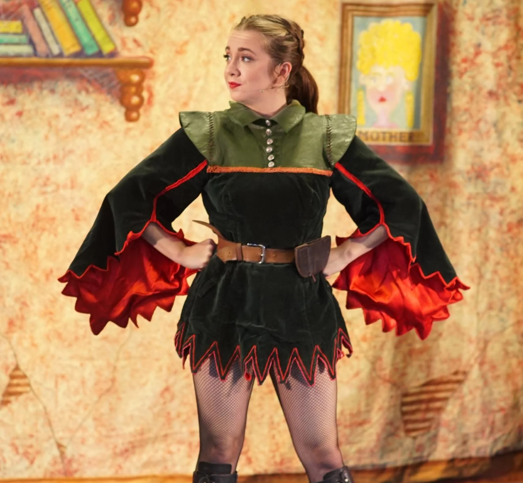 A young woman with light brown hair in a playful costume, standing with her hands on her hips in front of a colorful background, with a painting of a woman with curly yellow hair on the wall behind her.