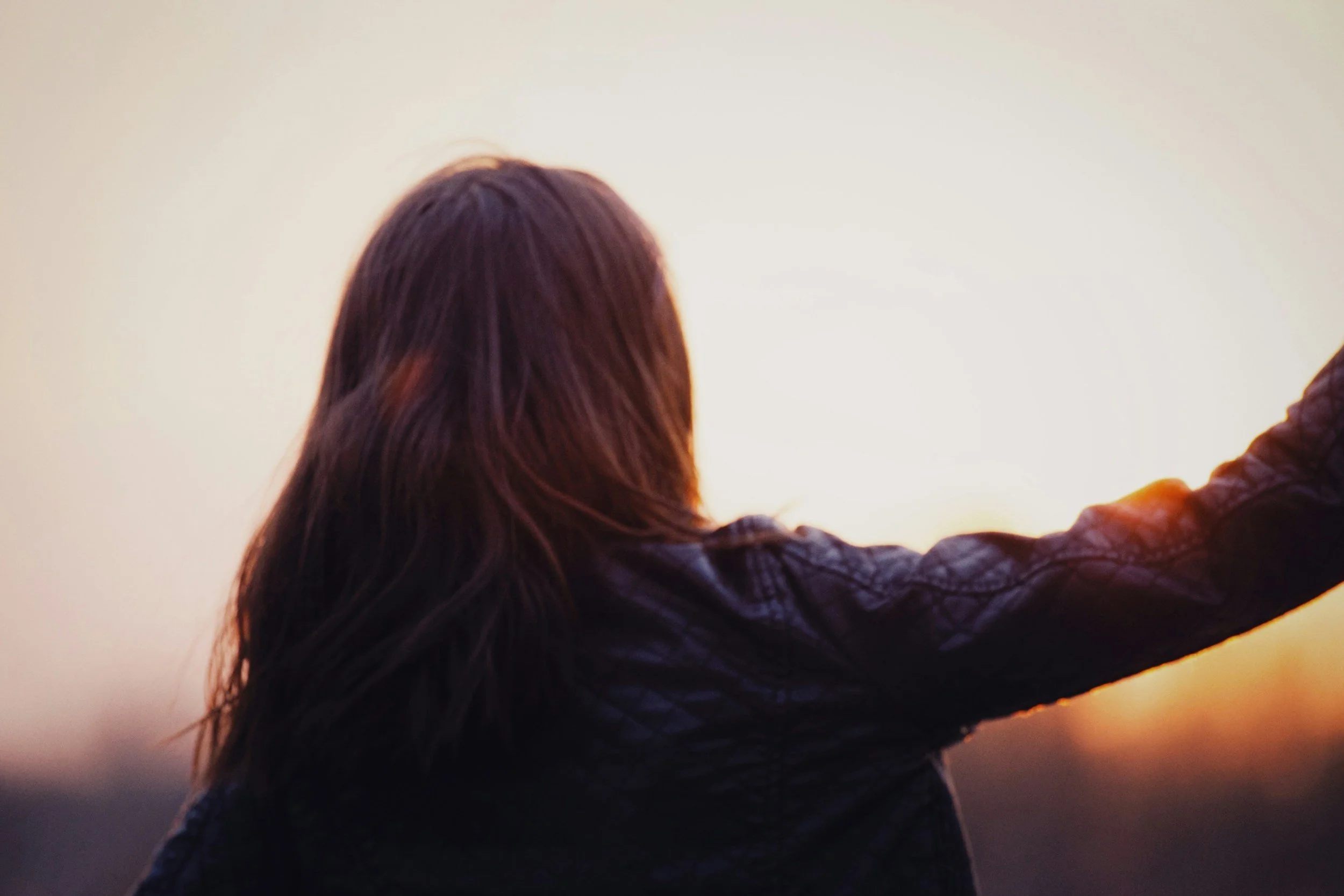 A person with long dark hair wearing a dark jacket, facing away, holding a camera or phone, capturing a sunset or sunrise over a body of water.