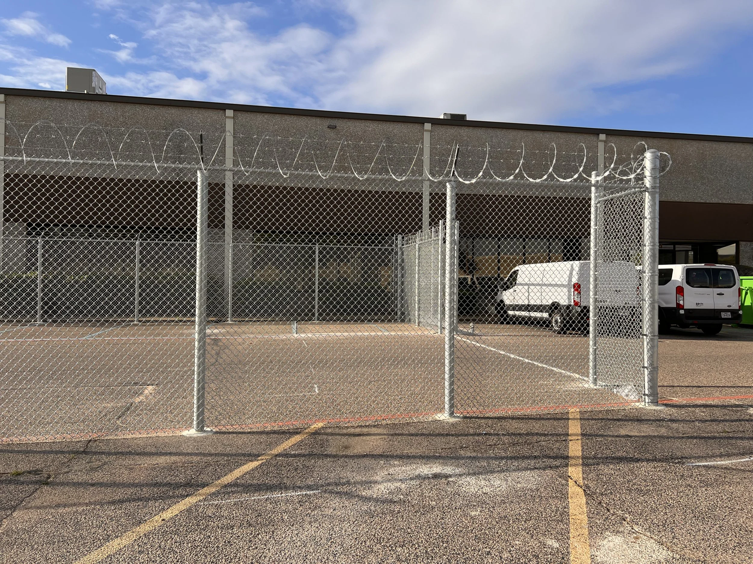 Fenced parking lot with white vans and a building in the background under a partly cloudy sky.
