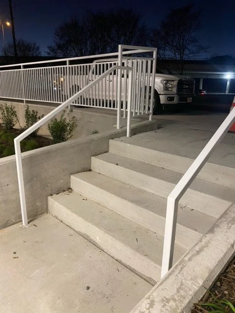 Concrete stairs with white metal railings leading up to a parking area with trucks in the background, at night.
