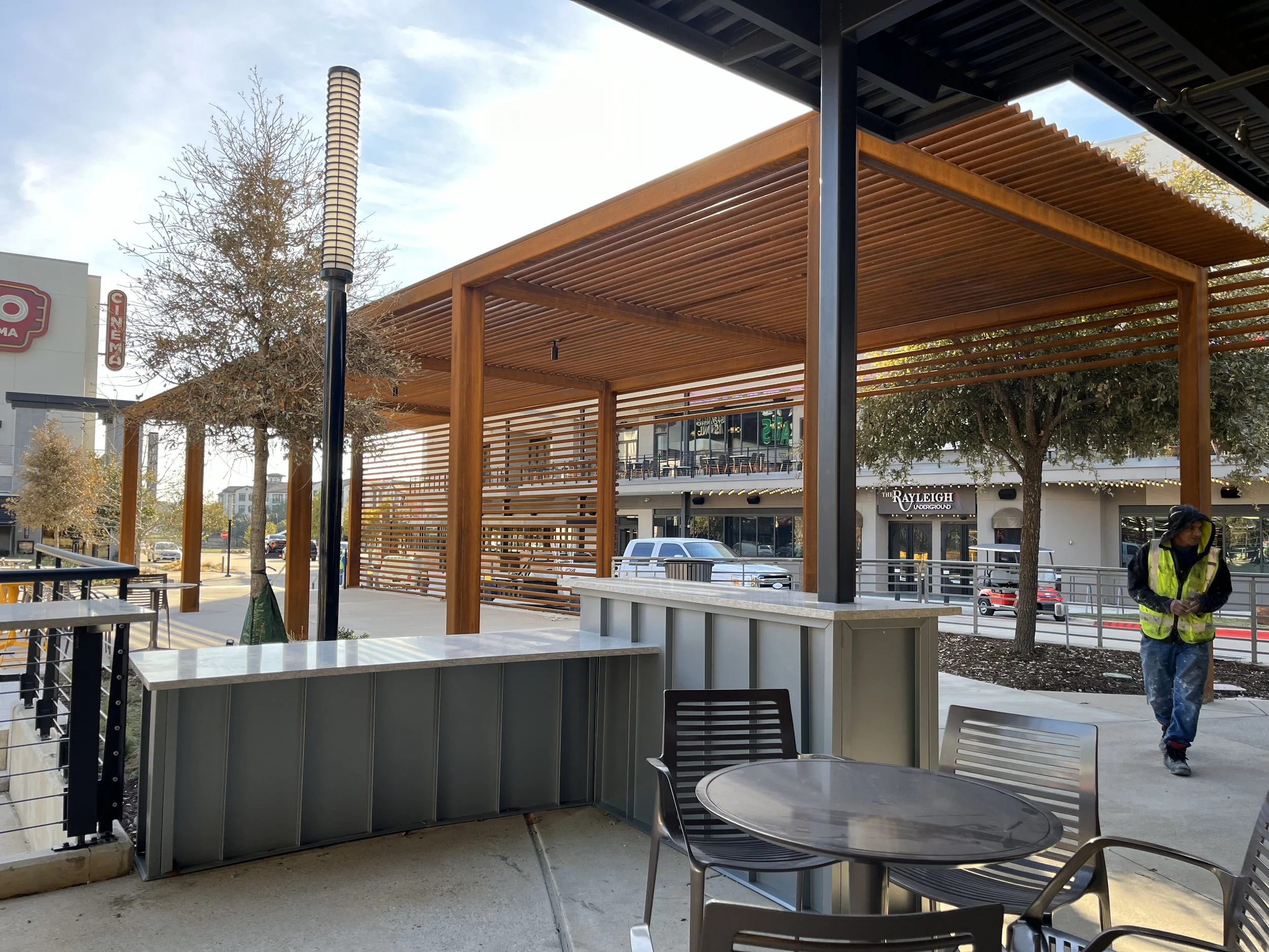 Outdoor patio with tables and chairs, pergola with wooden slats, a person in a yellow safety vest walking, trees, and buildings in the background.