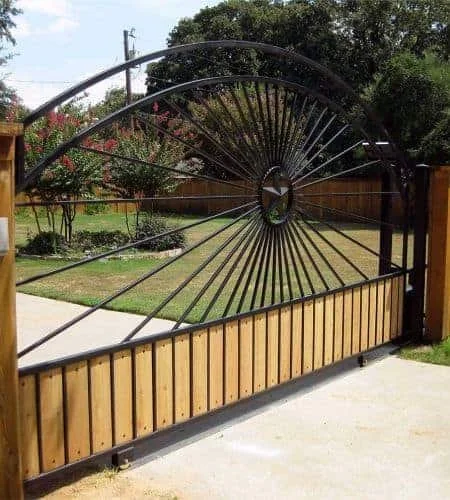 A decorative black metal gate with sunburst design, mounted on wooden posts, in a residential backyard with grass and trees.