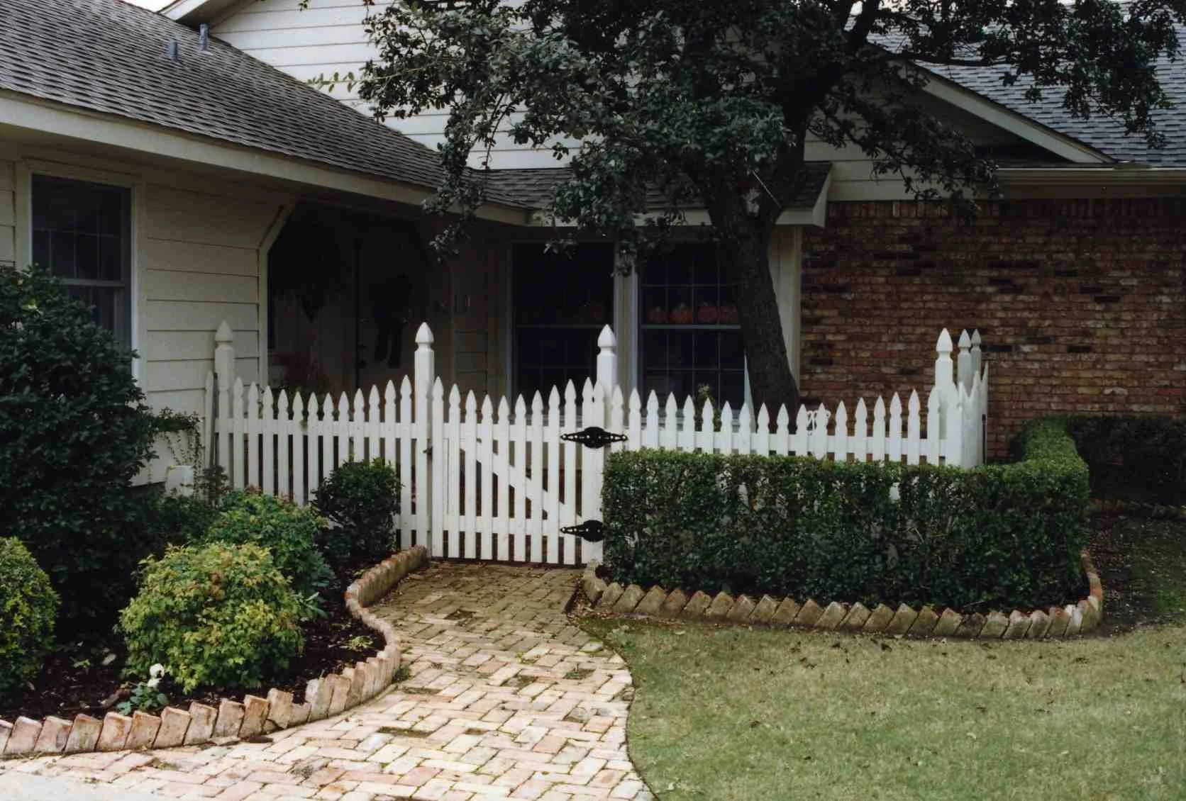 Front yard with a white picket fence, small brick pathway, green bushes, a large tree, and part of a house with brick and siding walls.