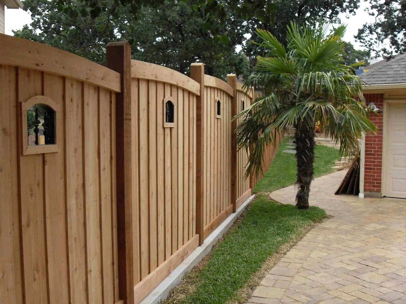 A wooden fence with small arched windows runs alongside a brick house with a driveway. There is a palm tree in front of the fence, and the area is landscaped with grass and a paved pathway.
