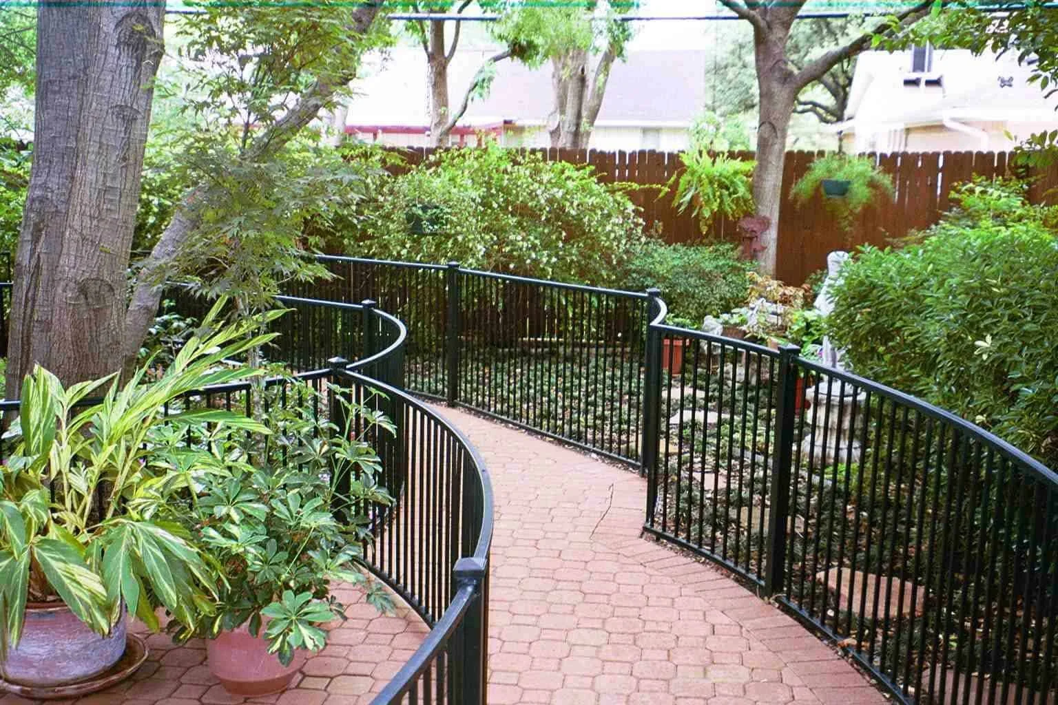 A winding brick pathway with black metal railings running along a lush garden with large trees, green shrubs, and potted plants, enclosed by a wooden fence.