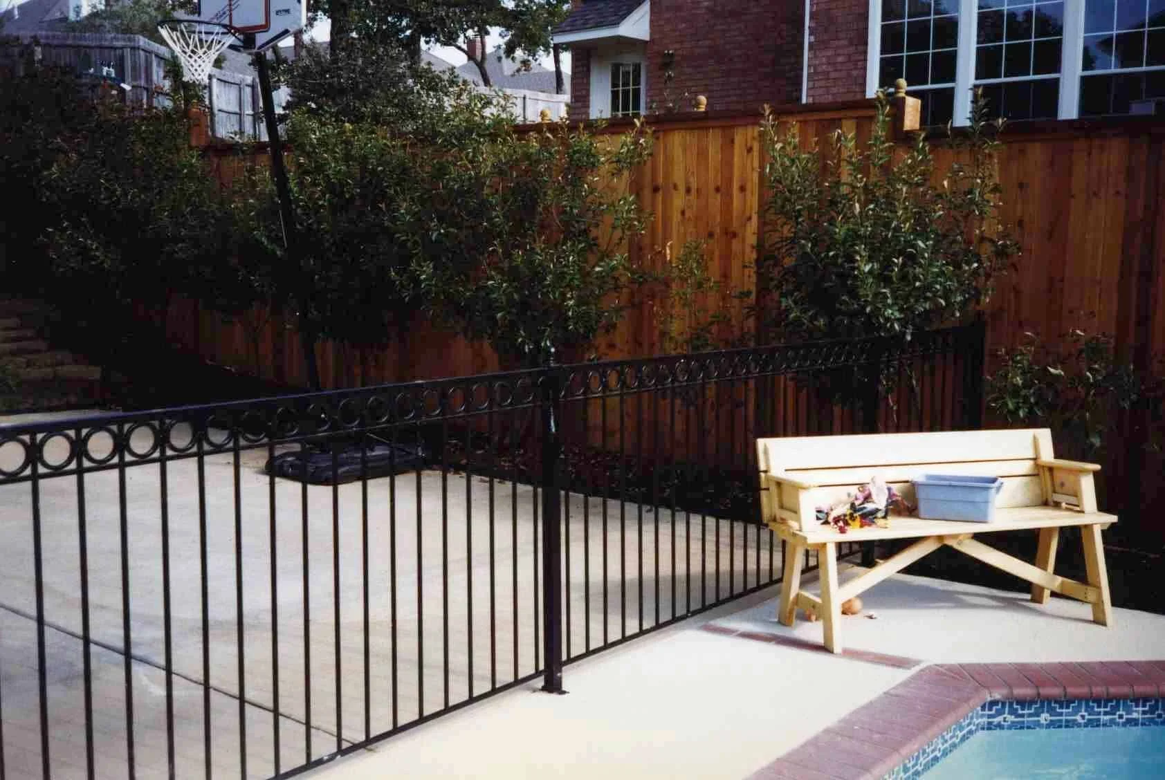 Backyard with a black metal safety fence, wooden bench, plastic box, and swimming pool, with trees, a brick house, and a wooden fence in the background.