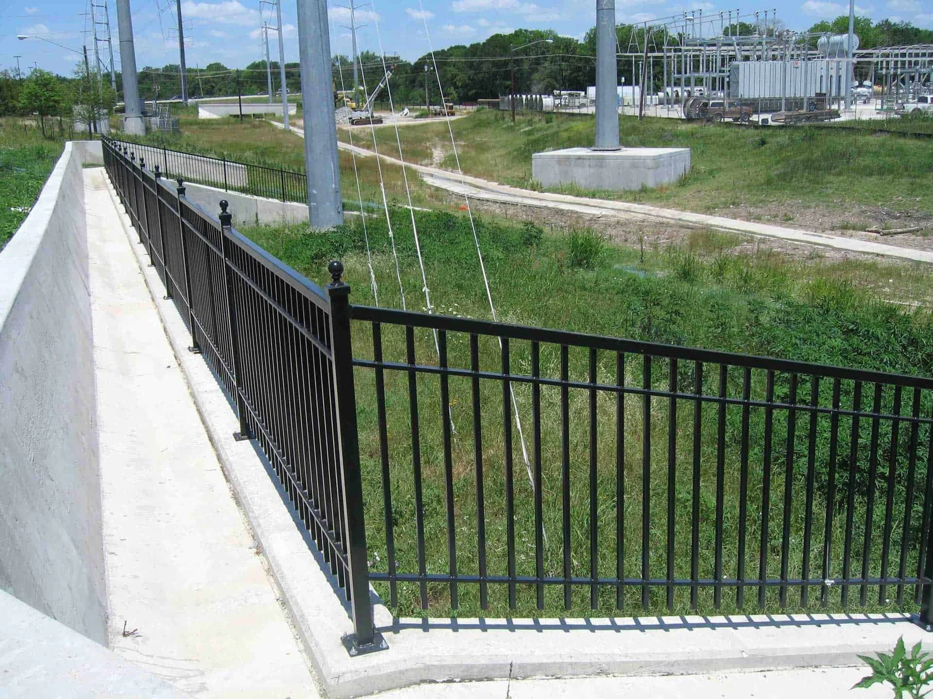 Black metal fence along a concrete sidewalk with grass and electrical infrastructure behind.