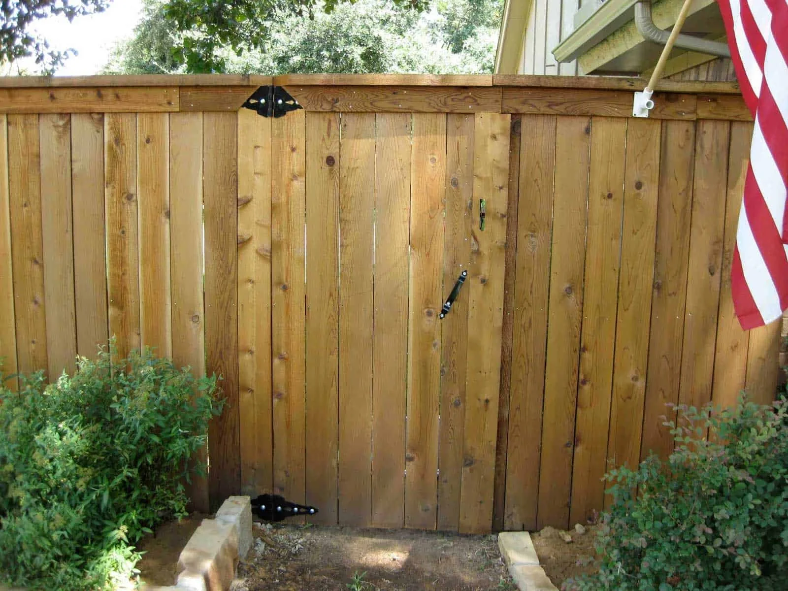 A wooden backyard fence with black metal hinges, a partially installed latch, and an American flag hanging on the right side.