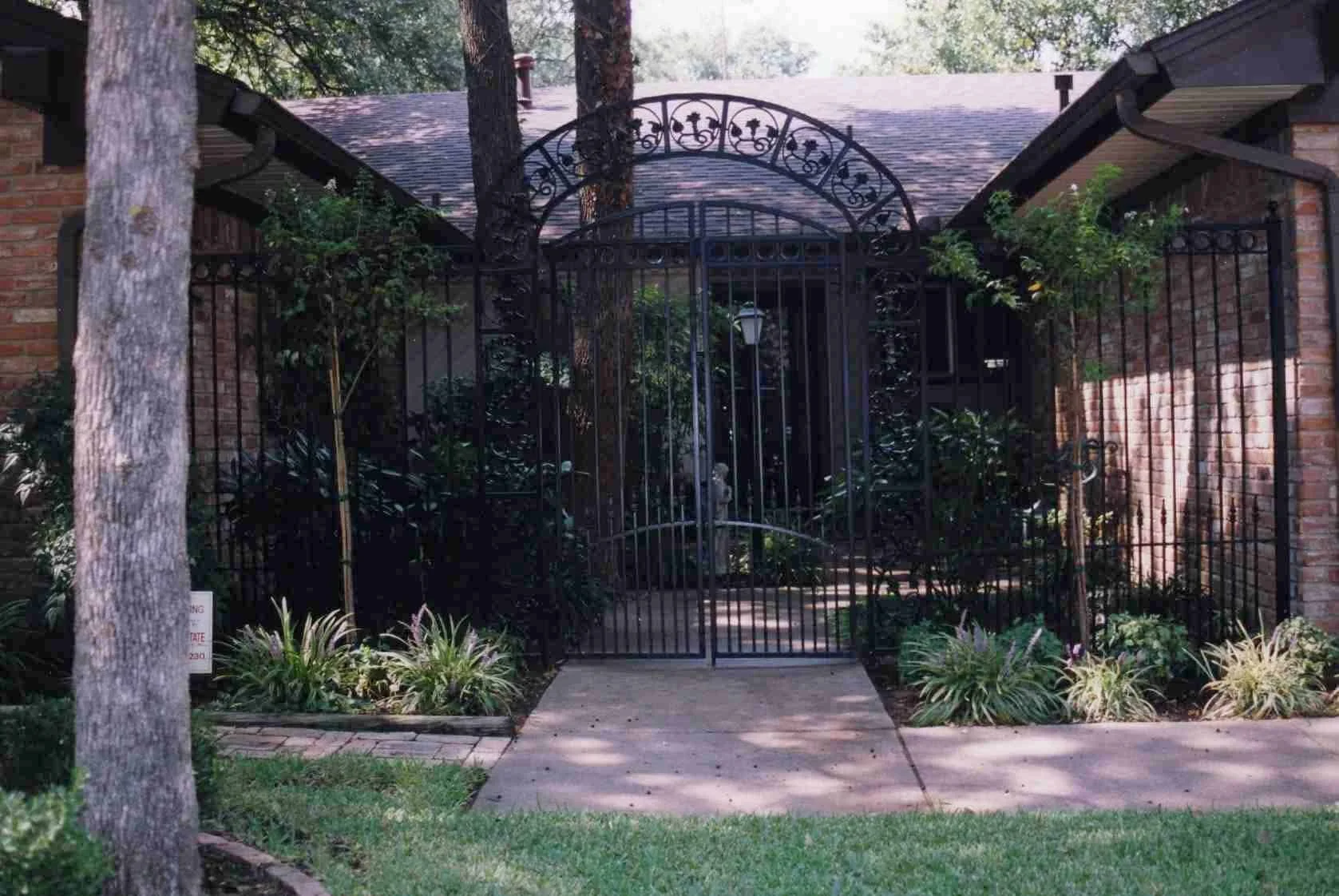 Black iron gate with decorative arch, surrounded by bushes and trees, leading to a brick house with a sloped roof.