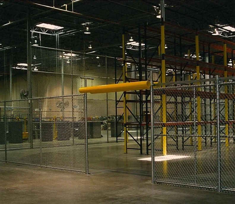 Inside a large industrial warehouse with metal shelving, yellow safety markings, and fencing, likely used for storage or manufacturing.