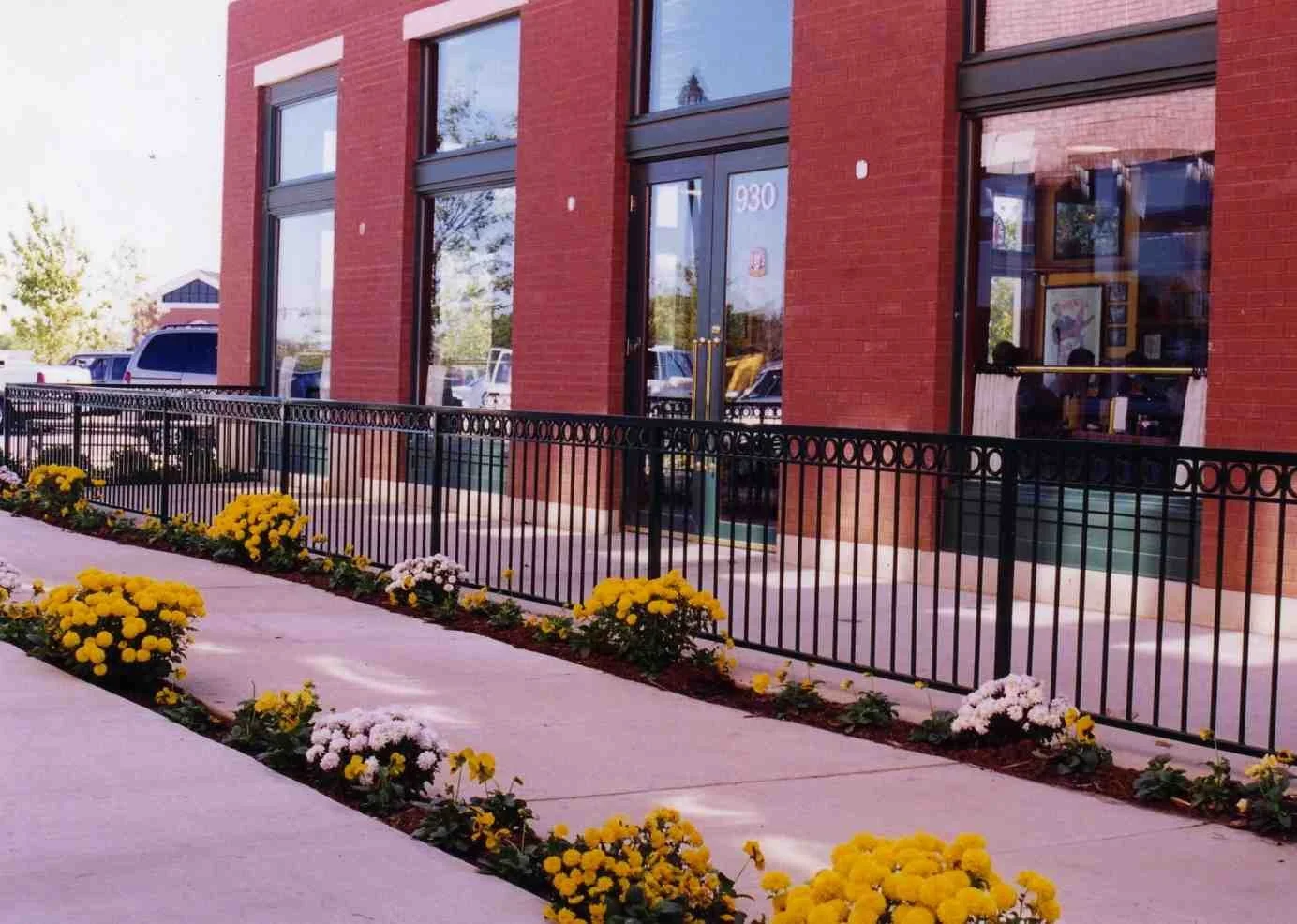 A cafe with large windows, red brick exterior, and black iron fence in front. There are flower beds with yellow and white flowers along the sidewalk.