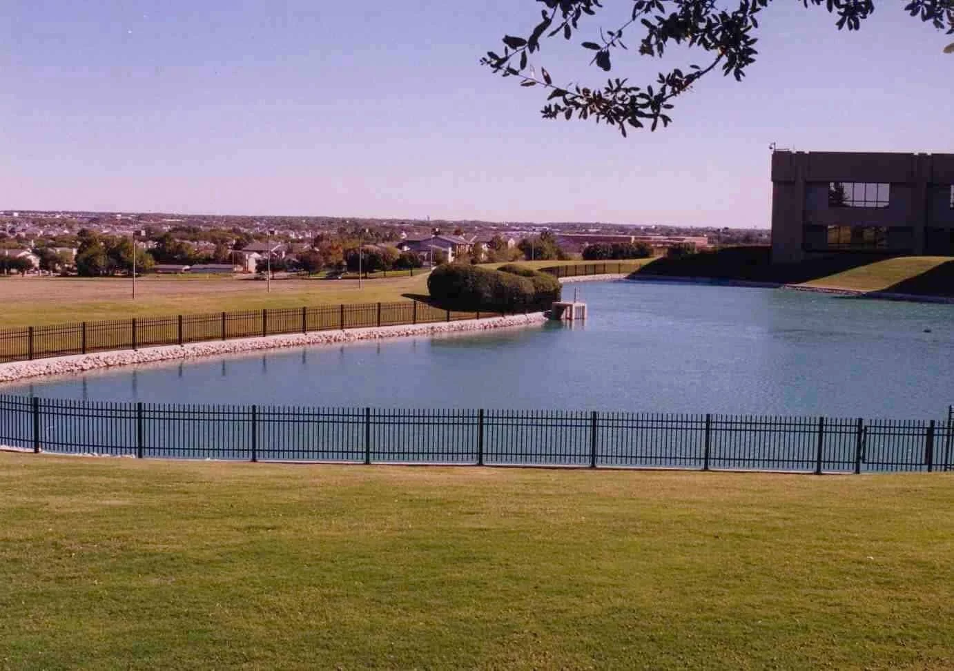 A body of water surrounded by a black fence with green grass in the foreground and a park with trees and buildings in the background.