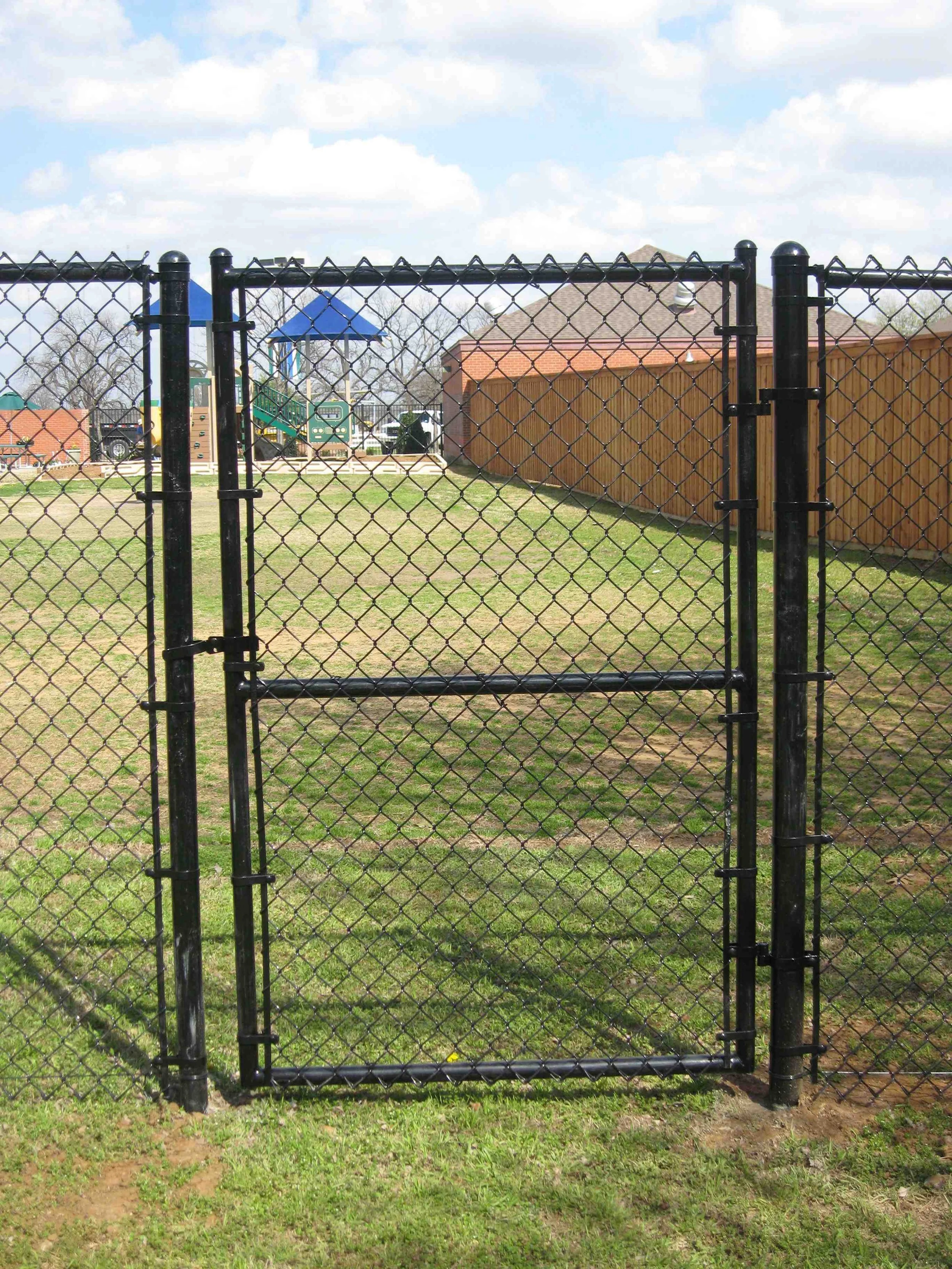 A black chain-link gate leading to a playground with a blue-roofed play structure, a small slide, and a fenced yard on a partly cloudy day.
