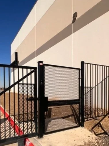 Black metal gate and fence beside a beige building wall under a clear blue sky.