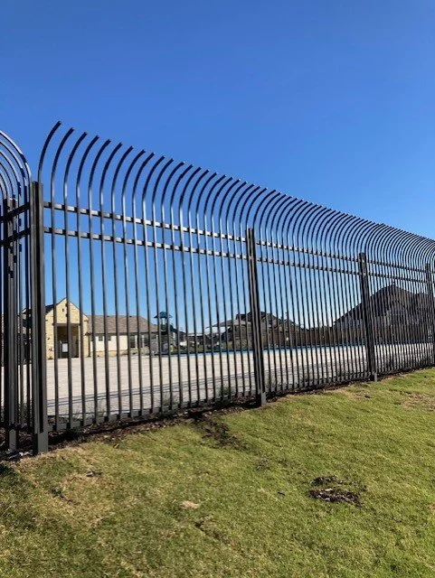 Tall black metal security fence with curved top spikes, in a suburban neighborhood with houses and a clear blue sky in the background.