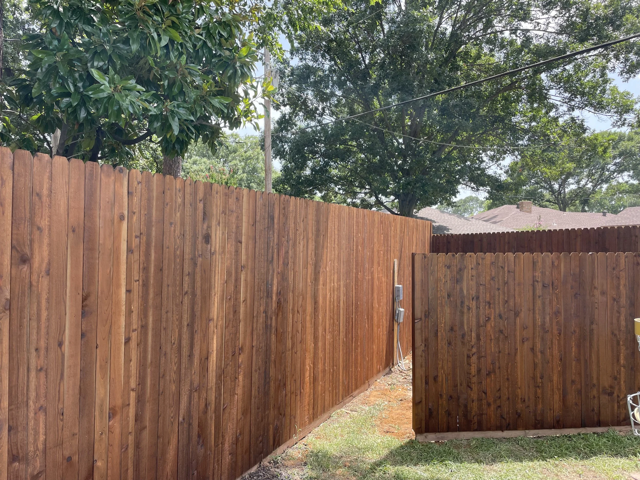 A backyard with two wooden fences, some trees in the background, and a partially visible chair on the right.