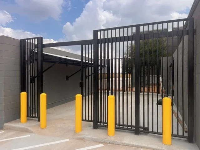 Black metal gate with yellow bollard posts blocking vehicle entry, leading to a parking area and covered section beyond, with a tree and building nearby under partly cloudy sky.