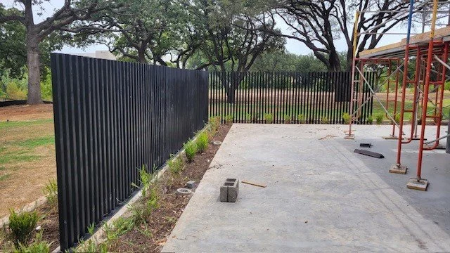 A black metal fence under construction, with a concrete slab and scaffolding, adjacent to some trees.