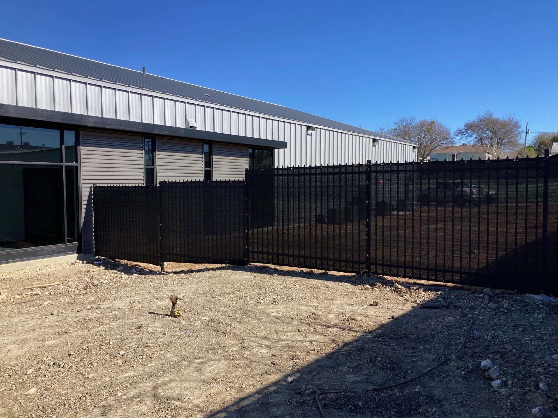 Construction site with a black metal fence, dirt ground, and a large metal building with metal siding and windows in the background under a clear blue sky.