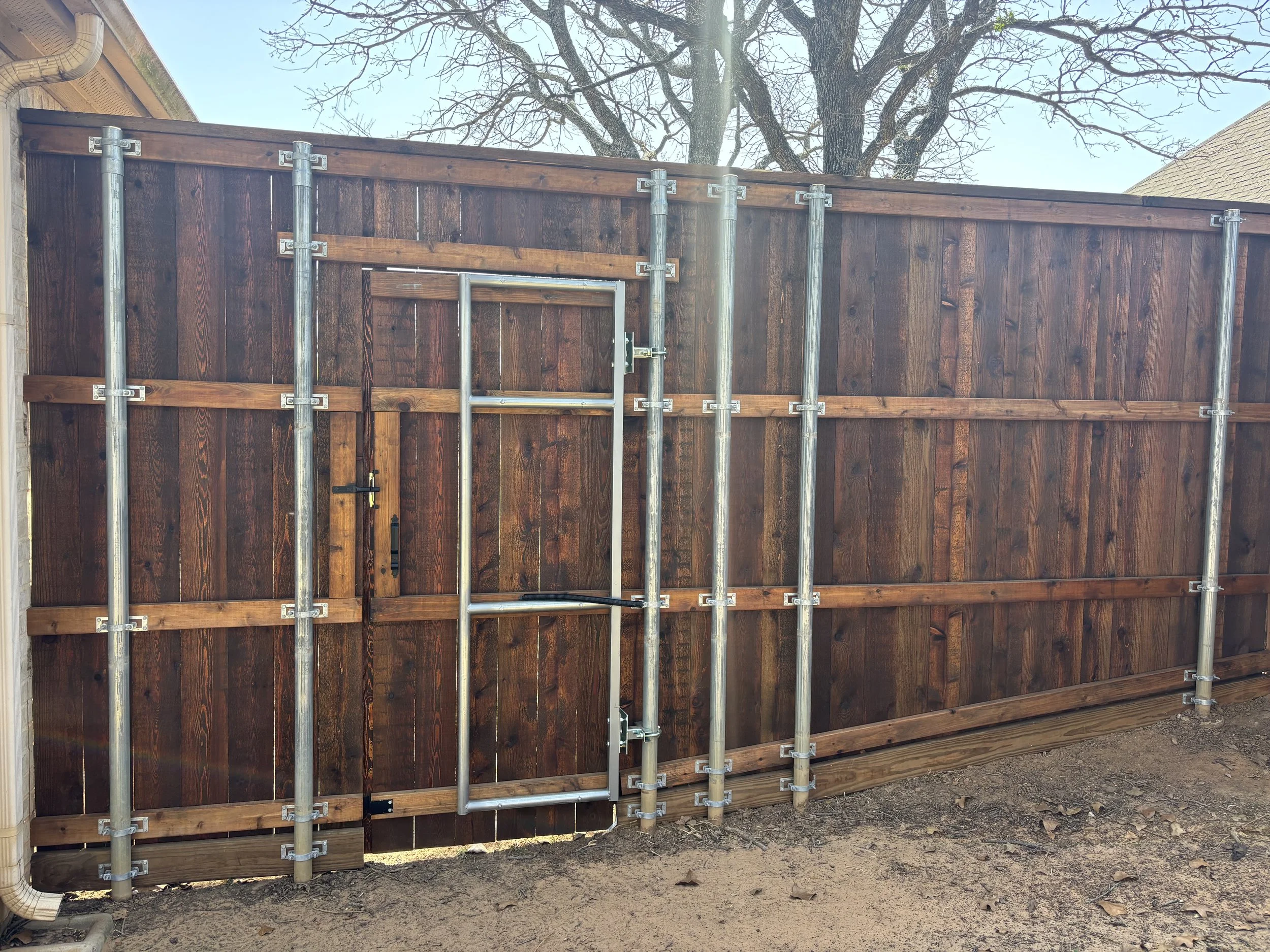 A wooden backyard fence with metal support poles, with a built-in metal dog door in the center, on a dirt ground outdoors.