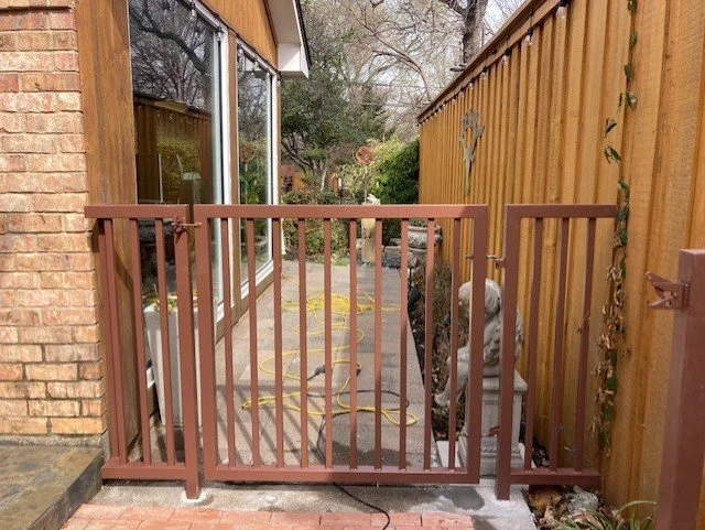 A small gated patio area between a brick house and a wooden fence, with a garden hose on the ground and some outdoor decorations.