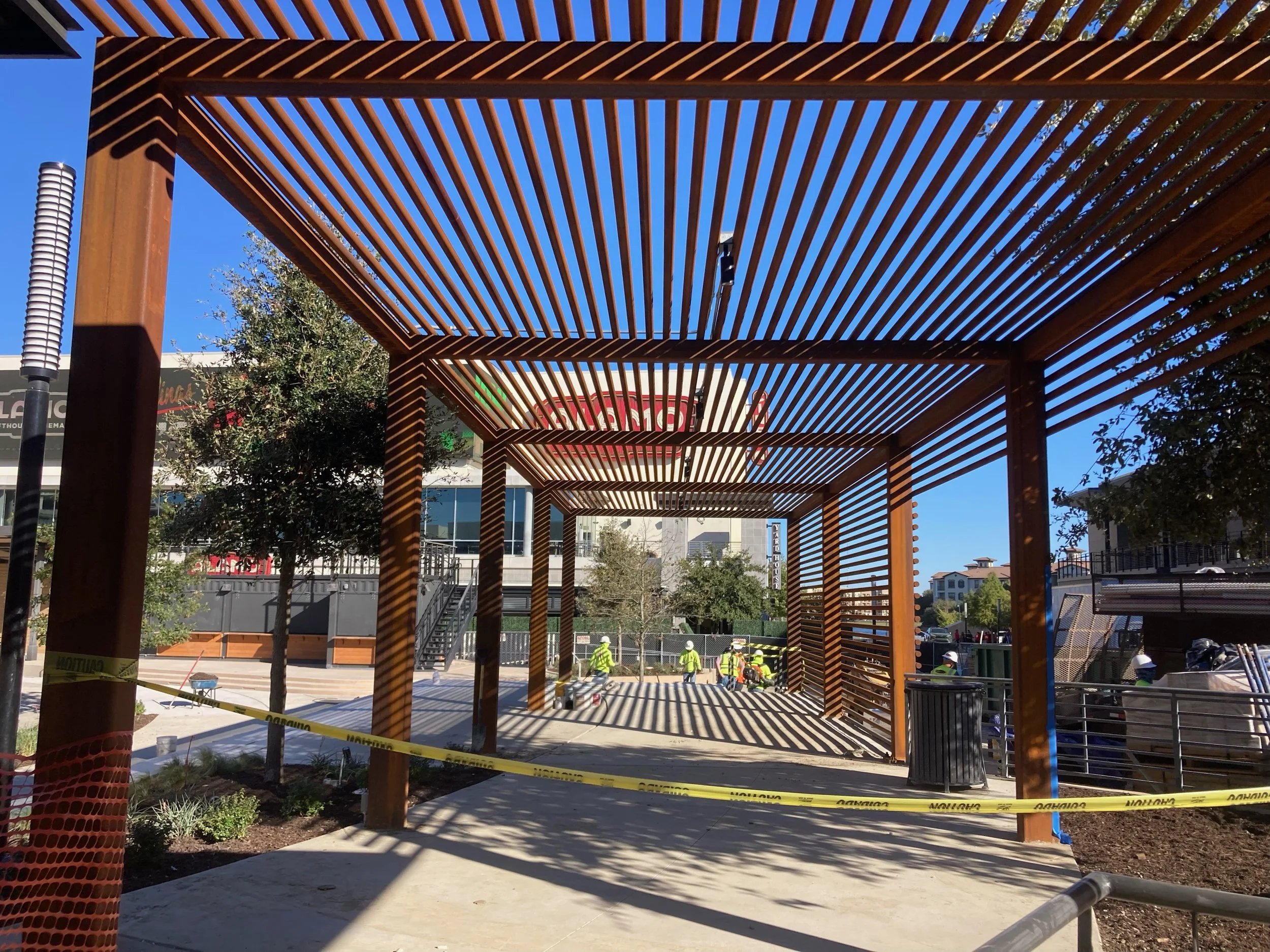 Wooden pergola with slatted roof casting striped shadows outside under clear blue sky, construction workers wearing yellow safety vests and helmets working nearby, caution tape surrounding the area.
