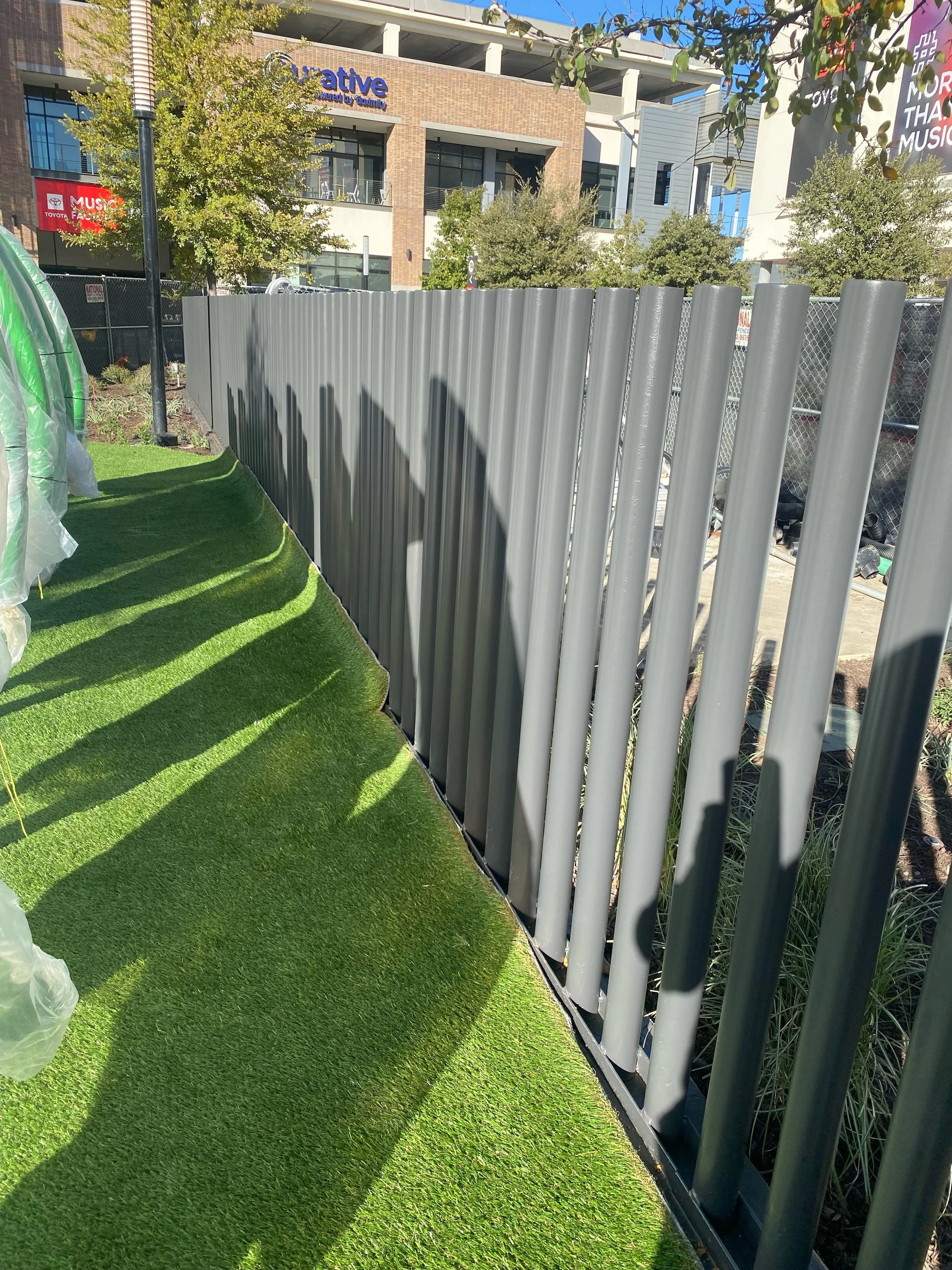 A view of a construction site that has a metal fence and a section of artificial grass on the ground, with the shadows of the fence cast on the grass. In the background, there are multi-story buildings with trees.