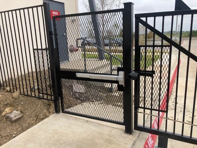 A black metal gate with a mesh pattern installed at an outdoor entrance, with a sidewalk in front and a fenced area behind, and some construction or landscaping work nearby.