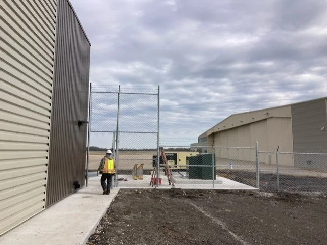 Construction worker walking near a chain-link fence next to industrial buildings under cloudy sky.