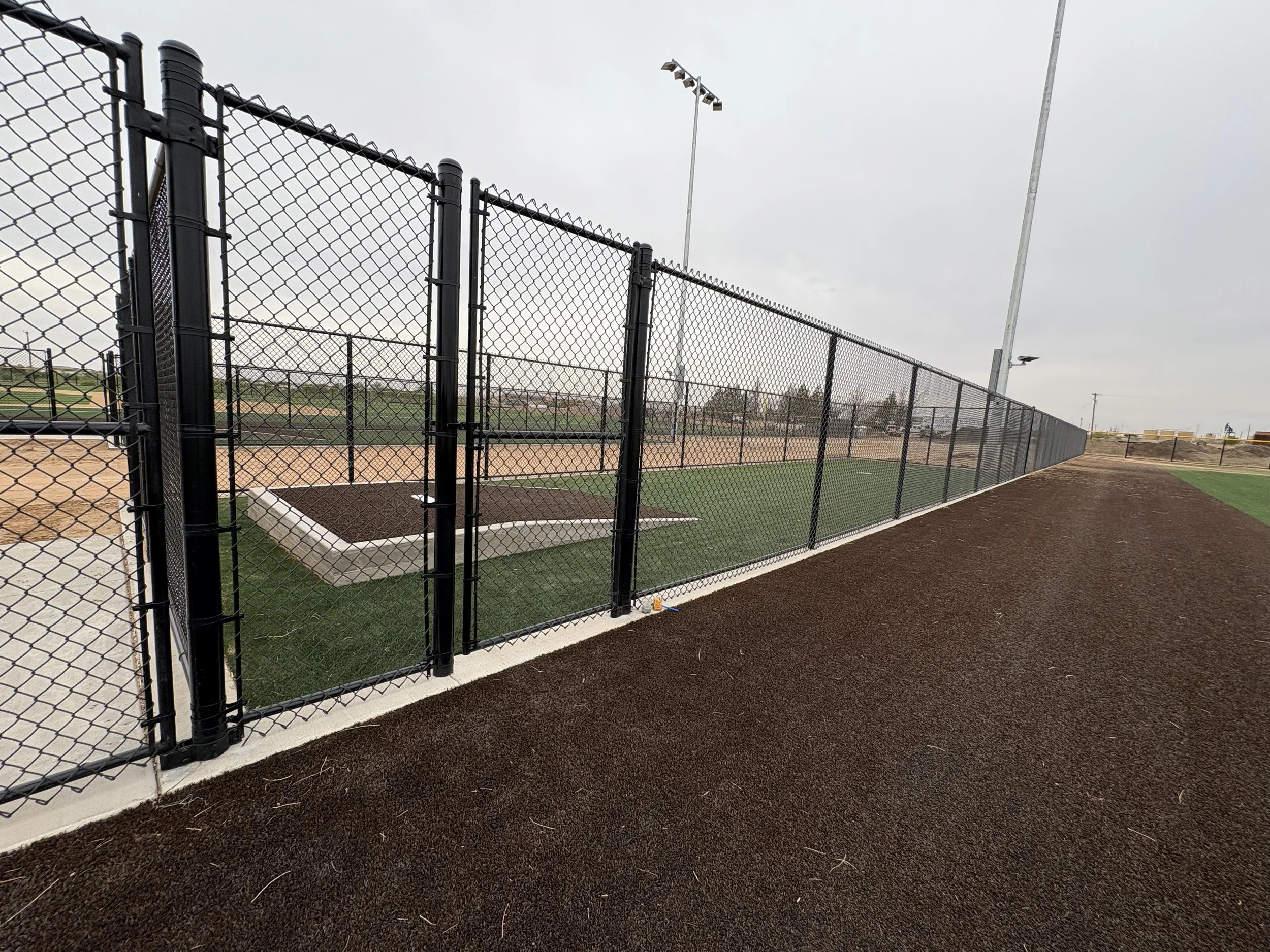 A baseball field viewed through a black chain-link fence with a dirt and grass infield, overcast sky, and light poles in the background.