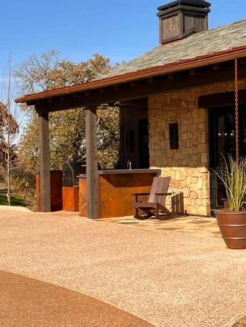 Outdoor patio area with stone house, wooden porch, and Adirondack chair, overlooking a backyard with trees.