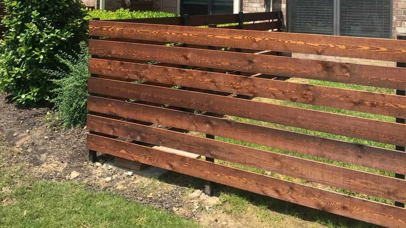 A wooden fence with horizontal slats in front of a house with plants and grass.