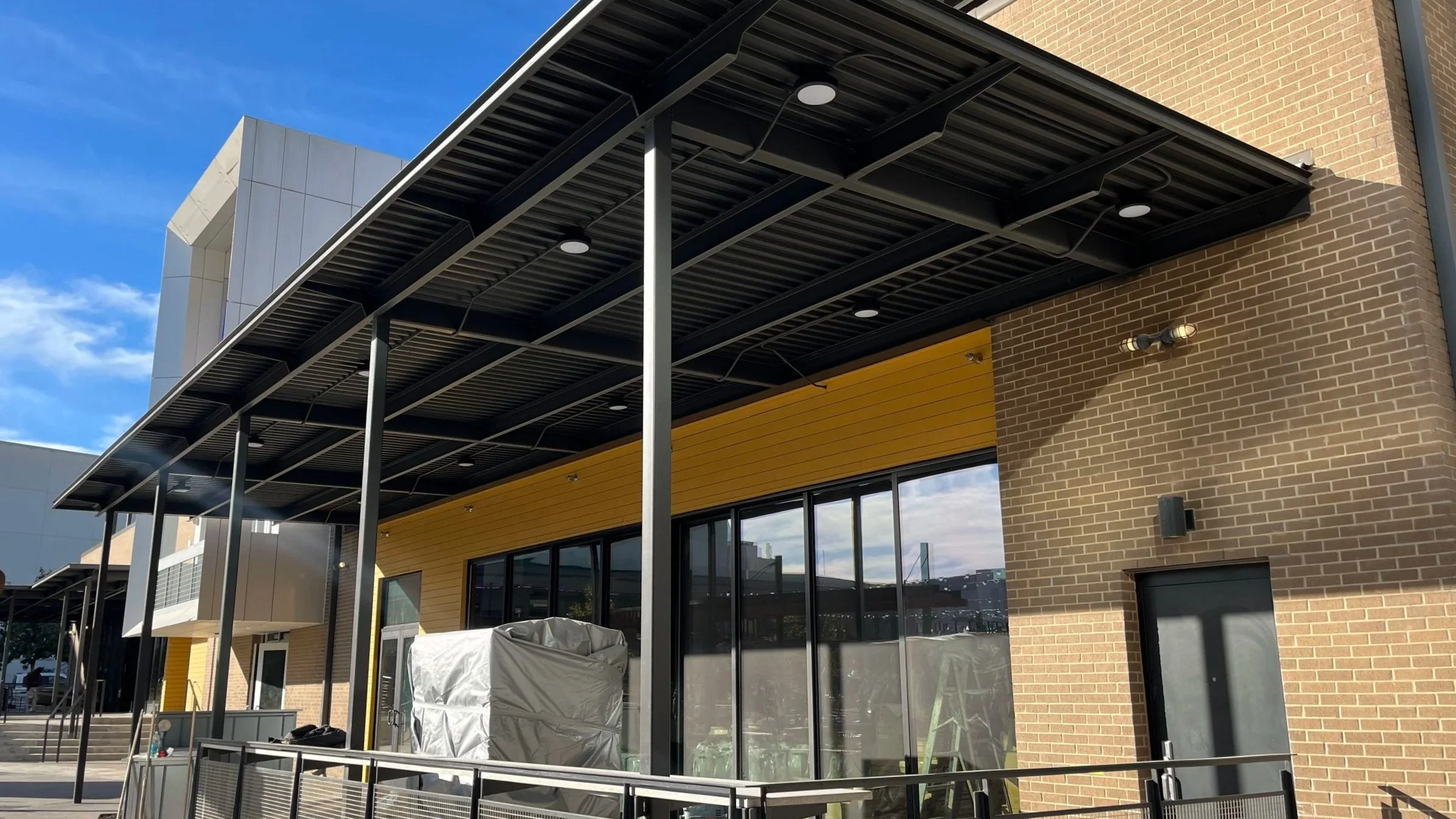 Modern commercial building with yellow and brick exterior, large glass windows, and a metal canopy overhead, under a partly cloudy blue sky.