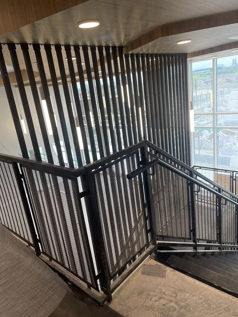 Indoor staircase with black metal railings, vertical wooden slats on a corner wall, large window showing a cityscape outside, and ceiling lights.