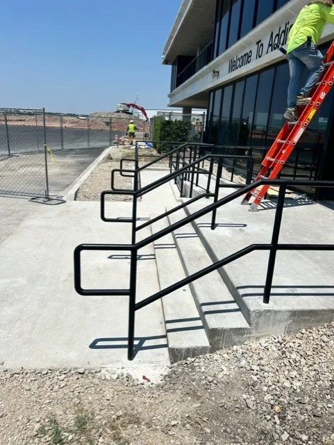 Construction workers installing black metal handrails outside a building with a sign partially visible that reads 'Welcome To Addi' and a red ladder leaning against the glass front.
