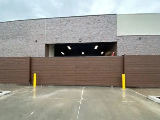 View of a parking lot with a brown wooden gate blocking access, yellow safety poles on either side, and a beige brick building in the background under a cloudy sky.