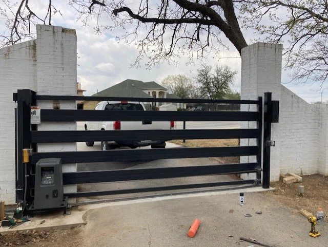 Motorized black security gate between white brick pillars with a pickup truck parked behind it, a tree above, and construction tools on the ground.