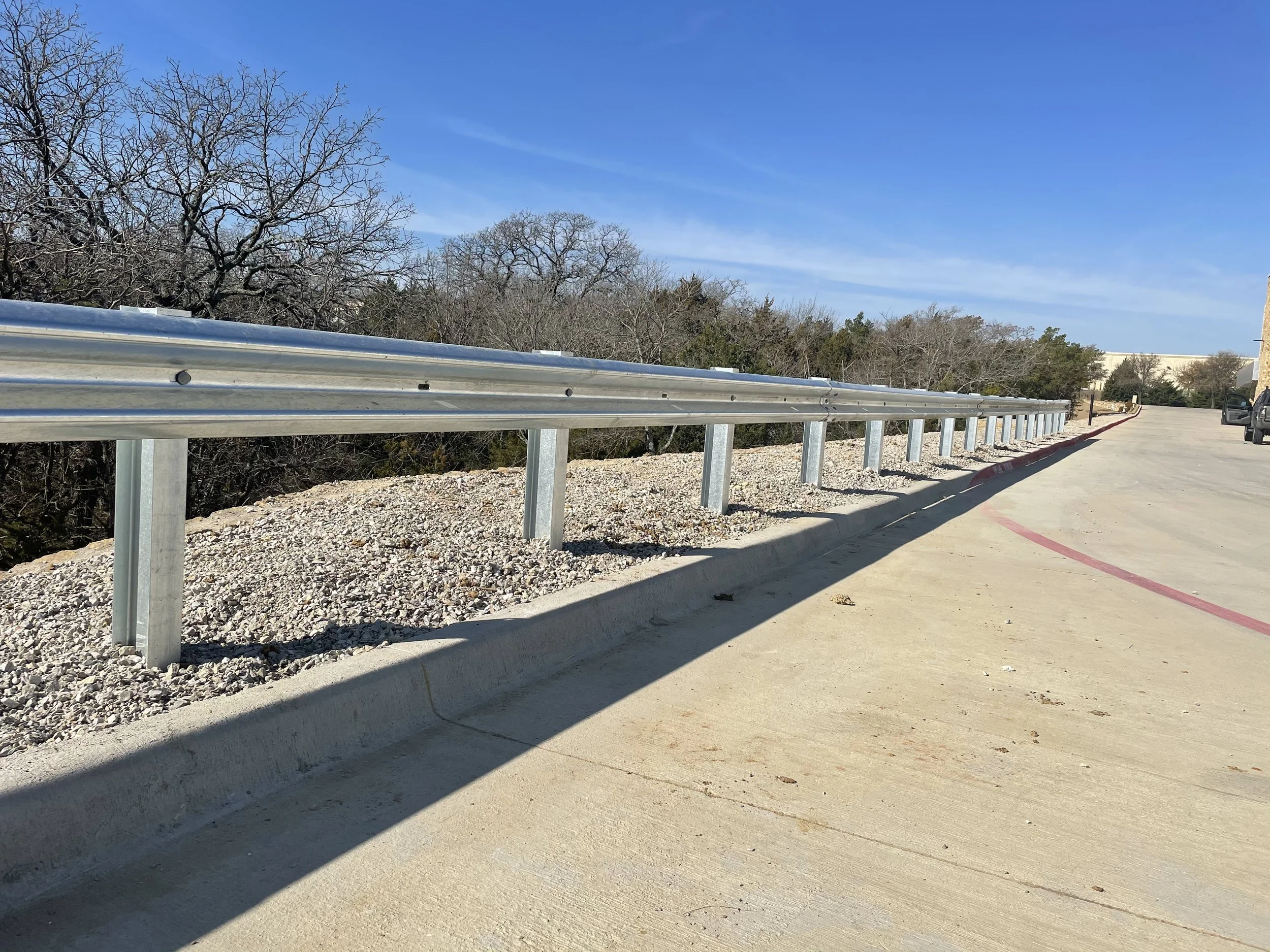 A concrete road with a metal guardrail on the side, casting a shadow on the pavement. There are trees with leafless branches and a building in the distance under a blue sky.