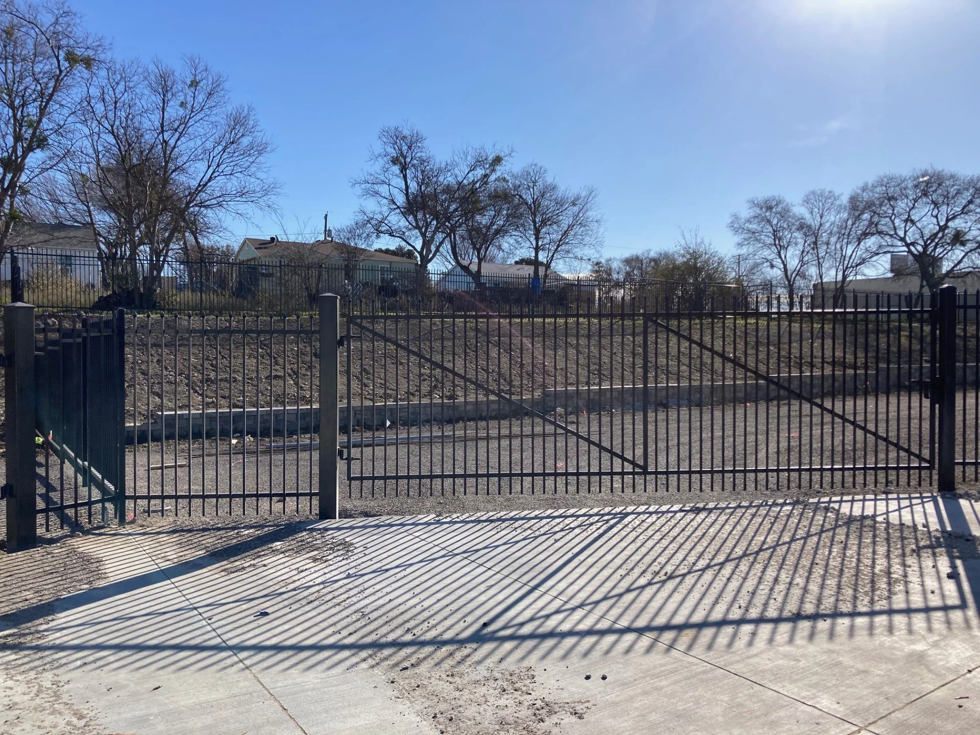 Empty black metal gate with shadows on concrete sidewalk, trees, houses, and clear sky in the background.