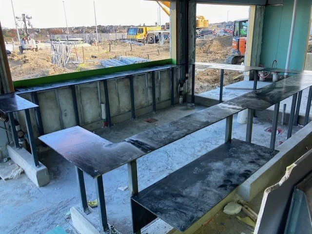 Construction site view from inside a building with a metal bar counter and a large window overlooking an active construction area with trucks and equipment.