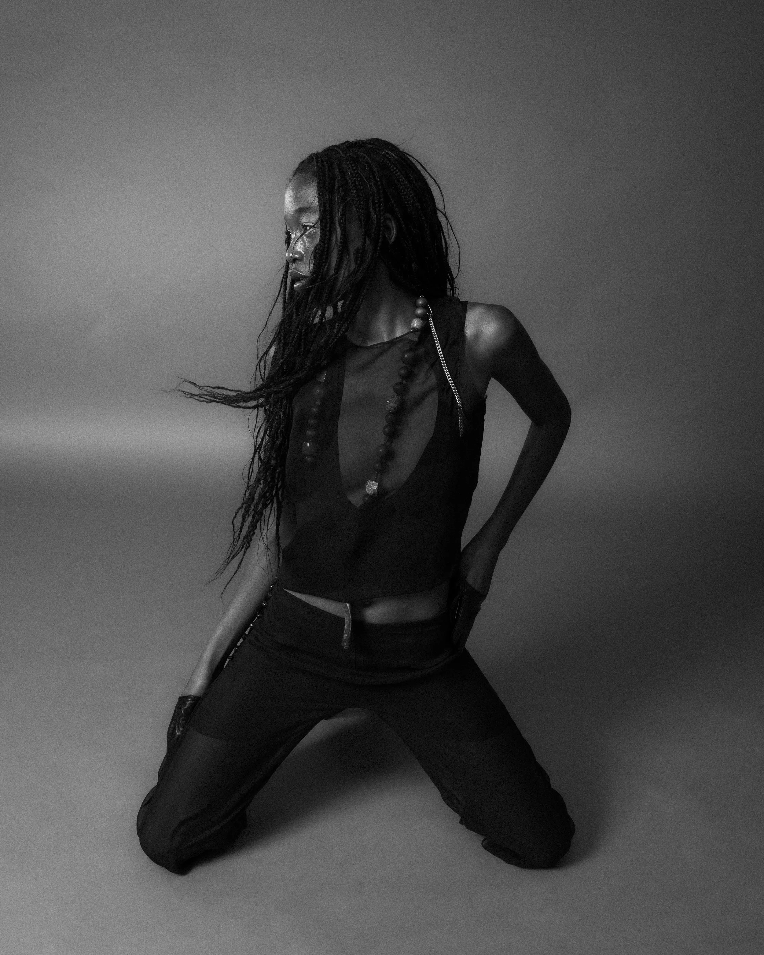 Black and white photo of a person kneeling on the floor, wearing a sleeveless top, long necklace, and pants, with long, flowing hair.