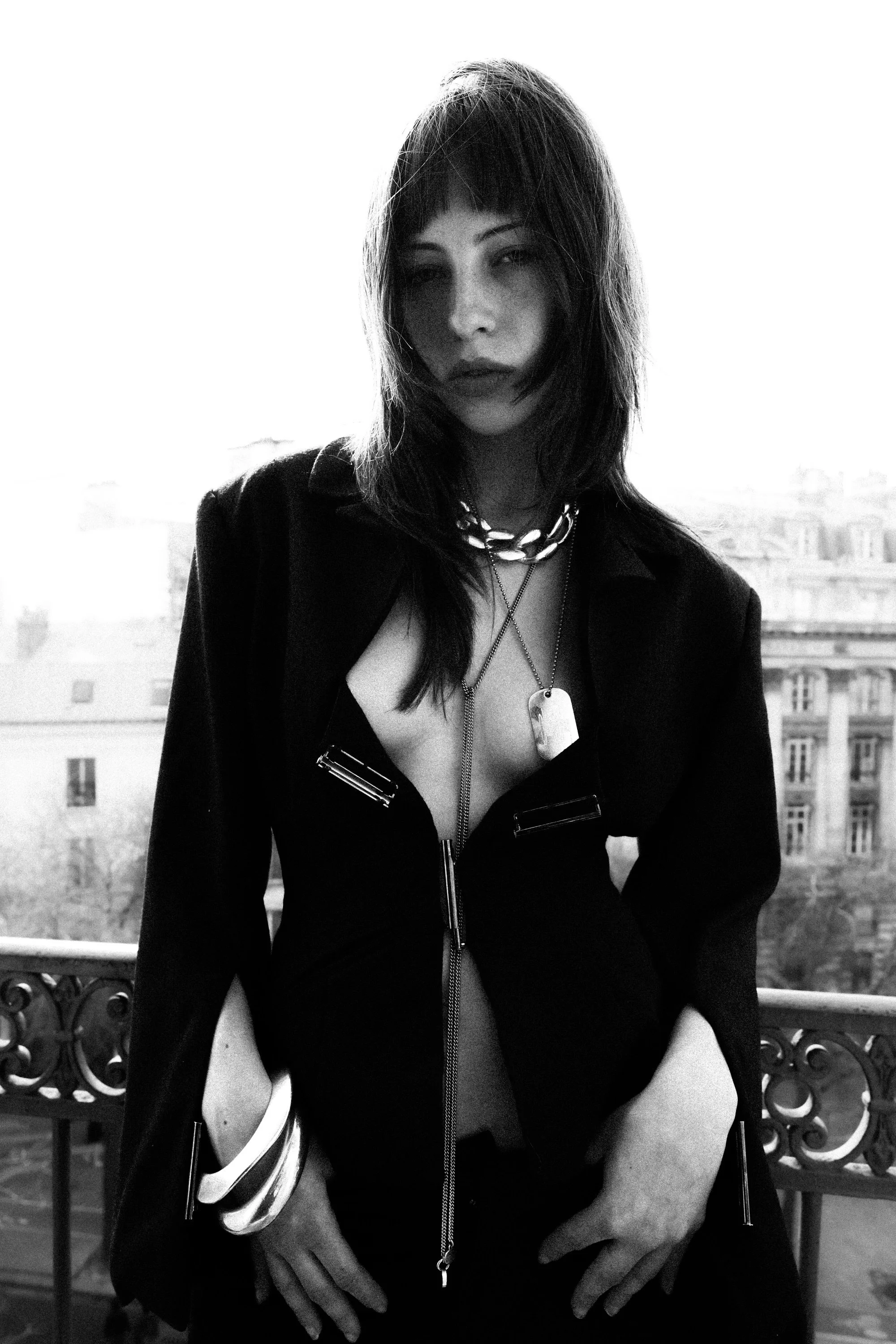 A woman with dark hair and a rebellious style posing on a balcony, dressed in a partially unzipped outfit with jewelry, with a cityscape in the background, in black and white.