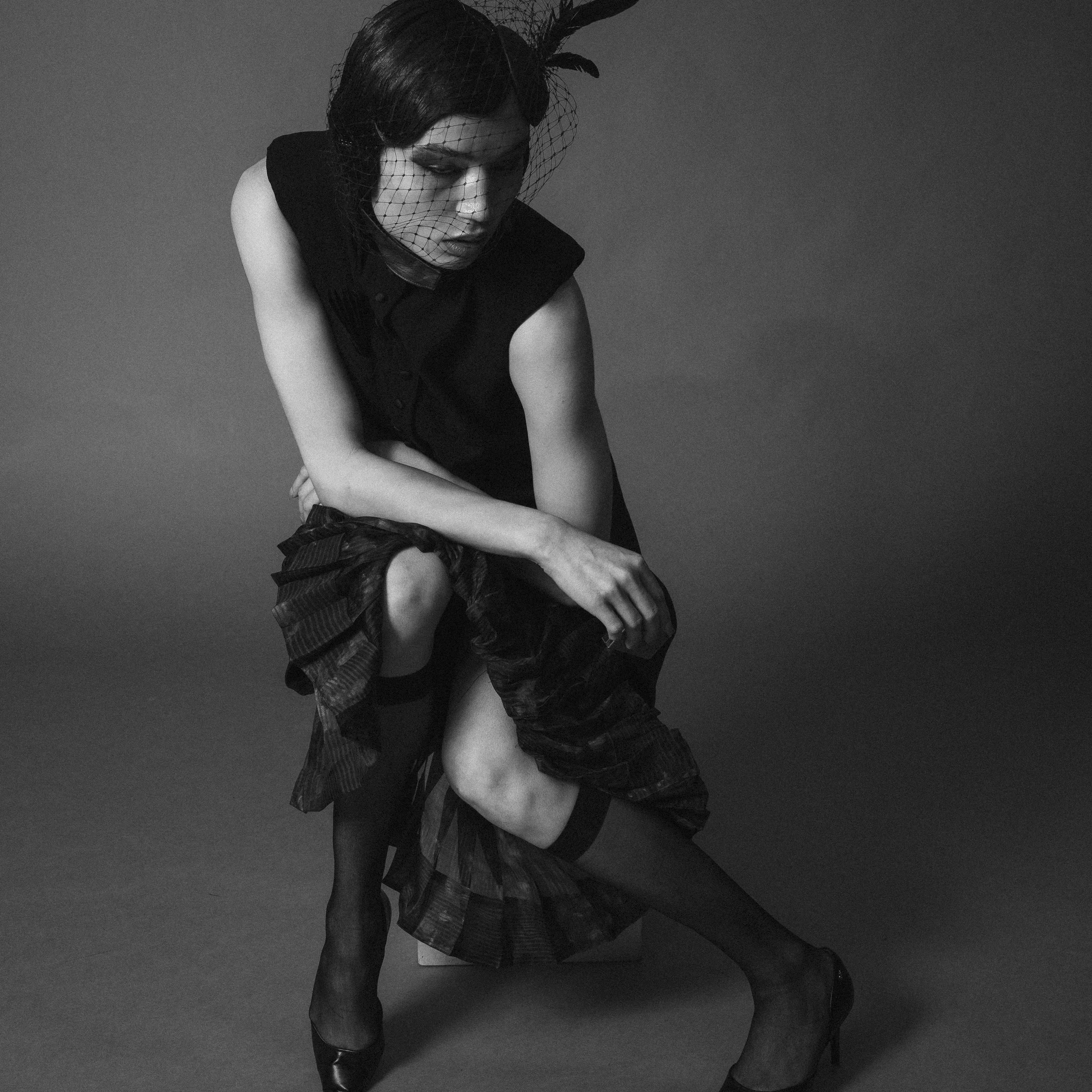 A woman with dark hair wearing a black dress, a netted headpiece, and thigh-high stockings, sitting and adjusting her shoe in a studio setting.