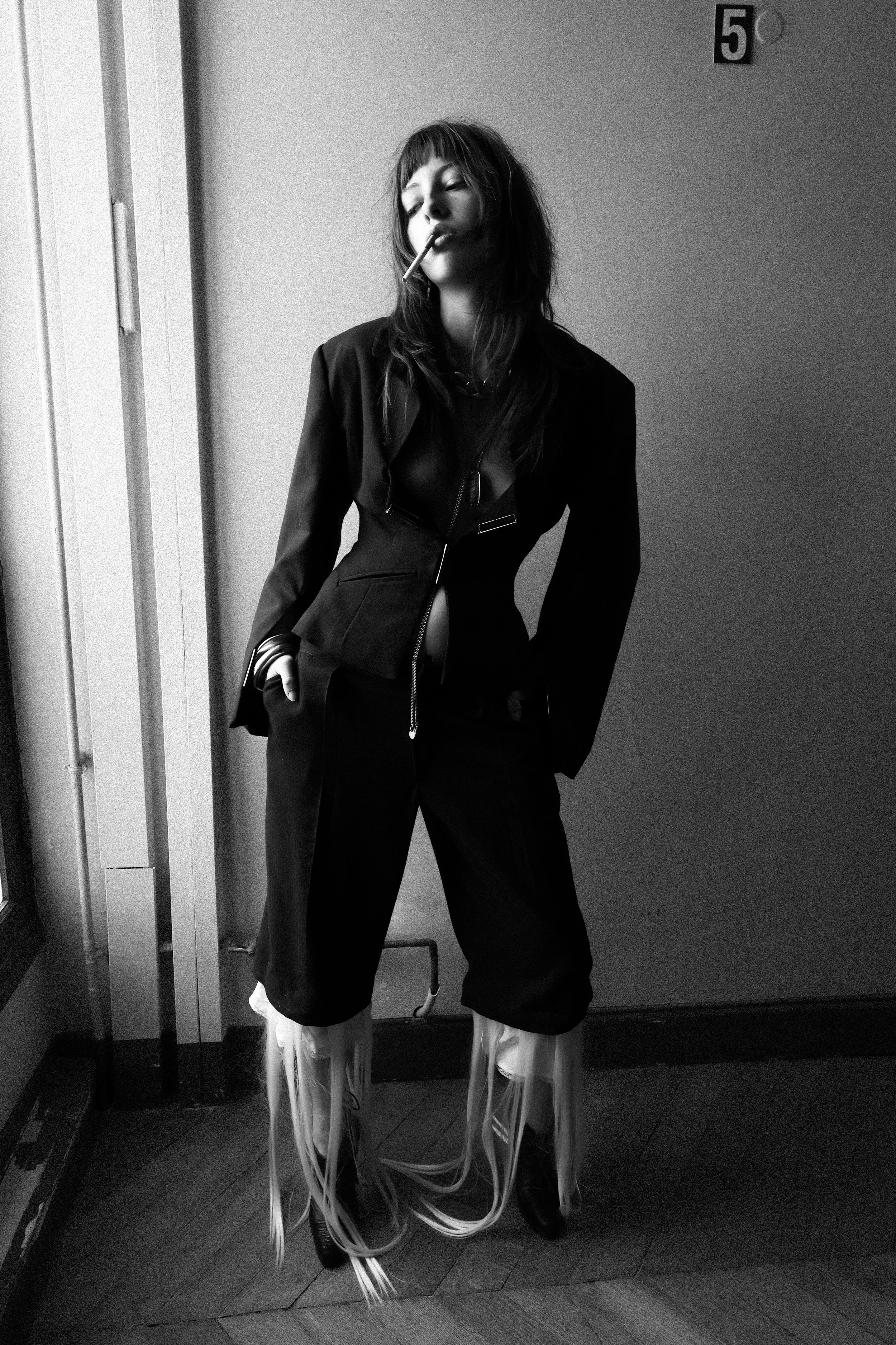 A woman with dark hair and bangs wearing a black blazer and pants with long, white fringe-like extensions on her legs, standing indoors against a plain wall with the number '5' above her head, smoking a cigarette.