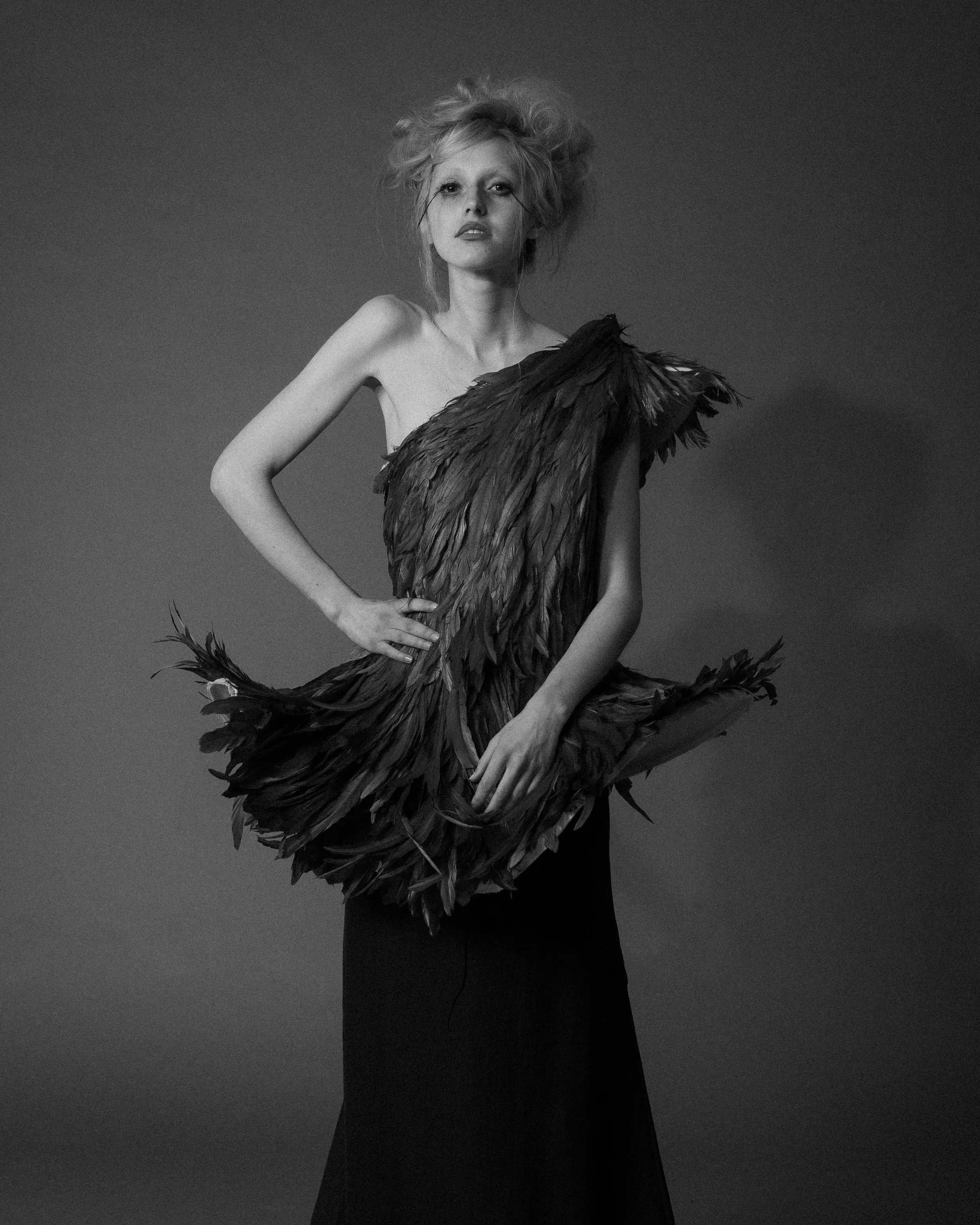 Black and white photo of a person wearing a dress with feather details, striking a pose against a plain backdrop.