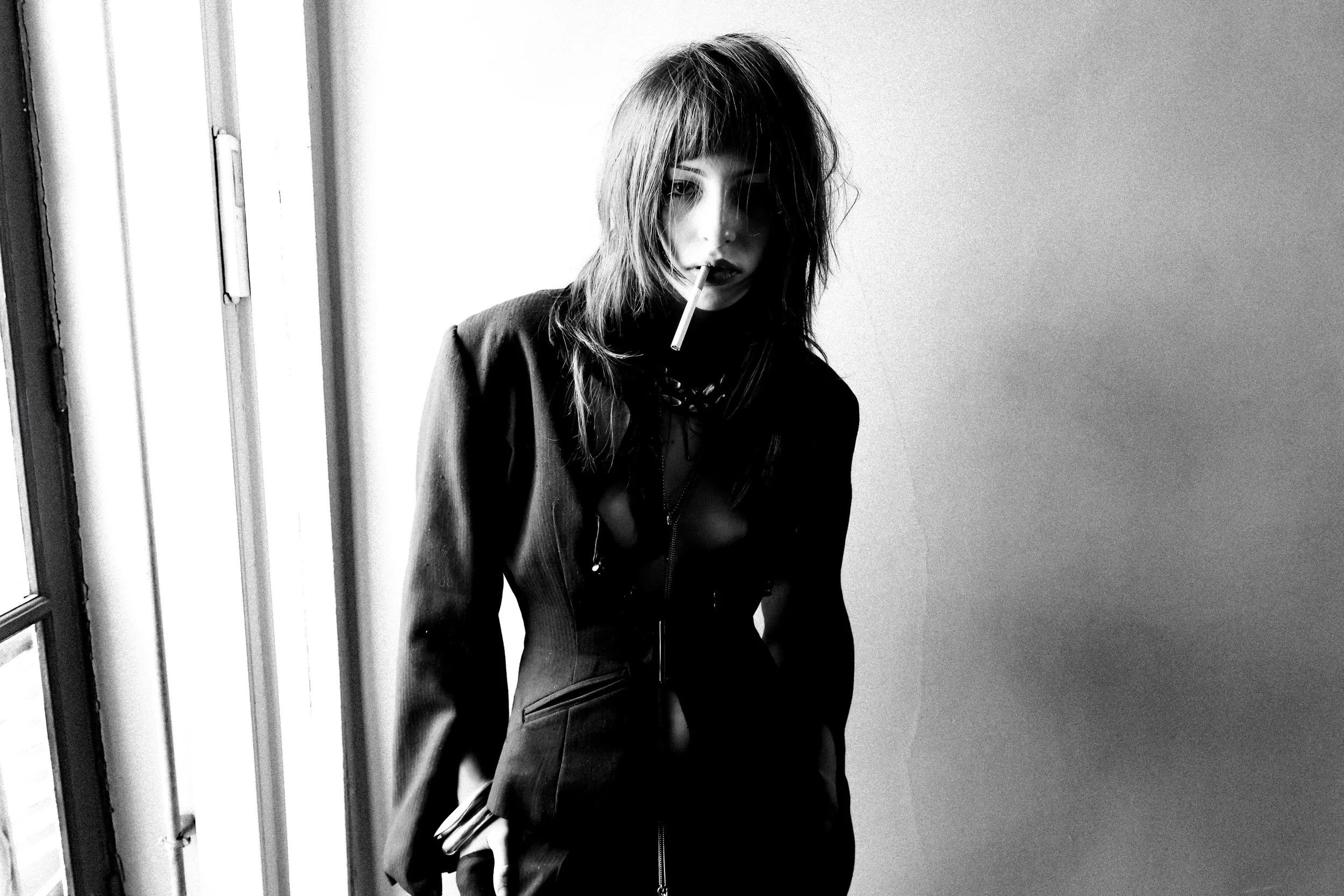 Black and white photo of a woman with shoulder-length hair, wearing a dark jacket, holding a cigarette in her mouth, standing near a window with a serious expression.