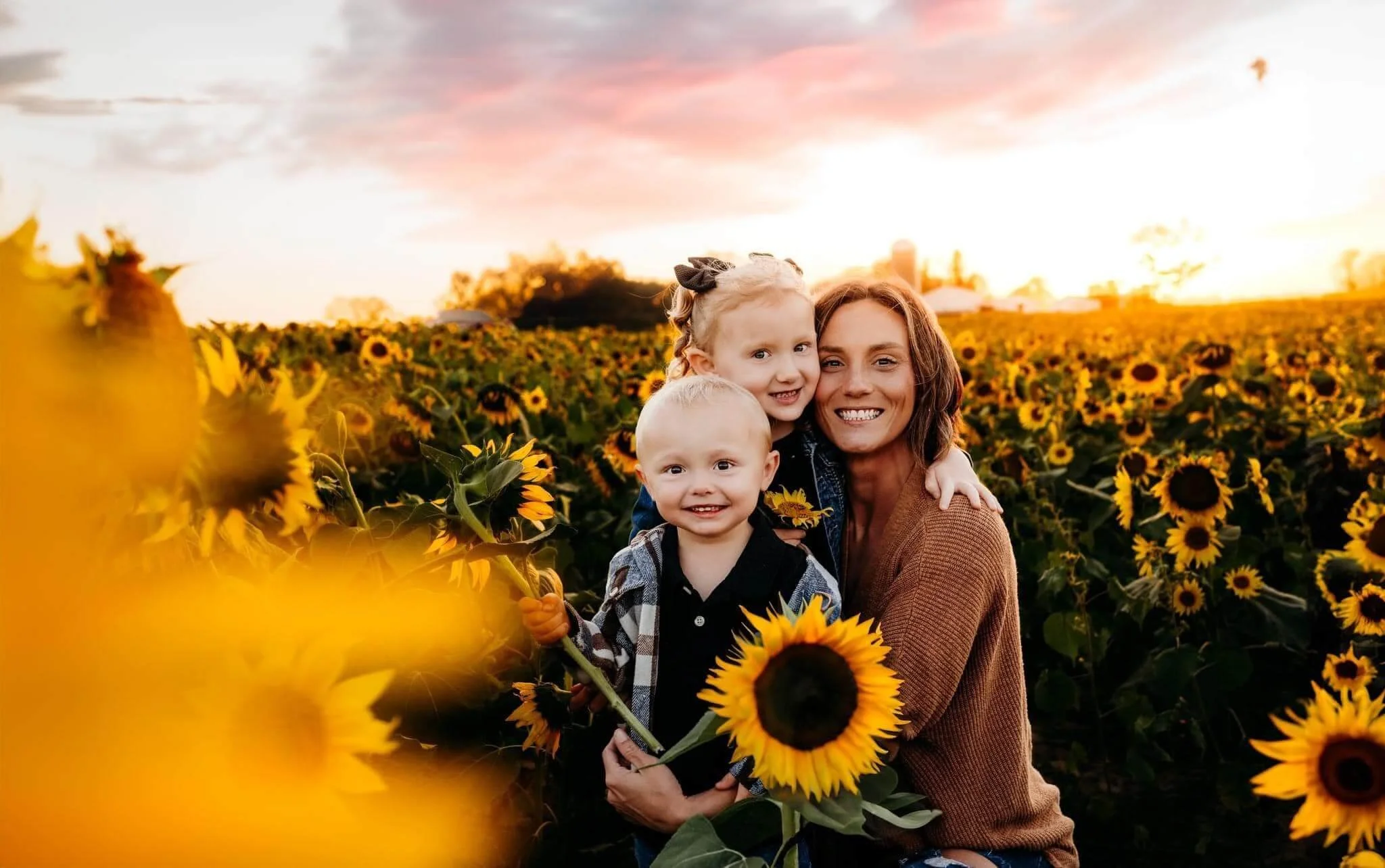 A woman and two children smile and hug in a sunflower field at sunset.