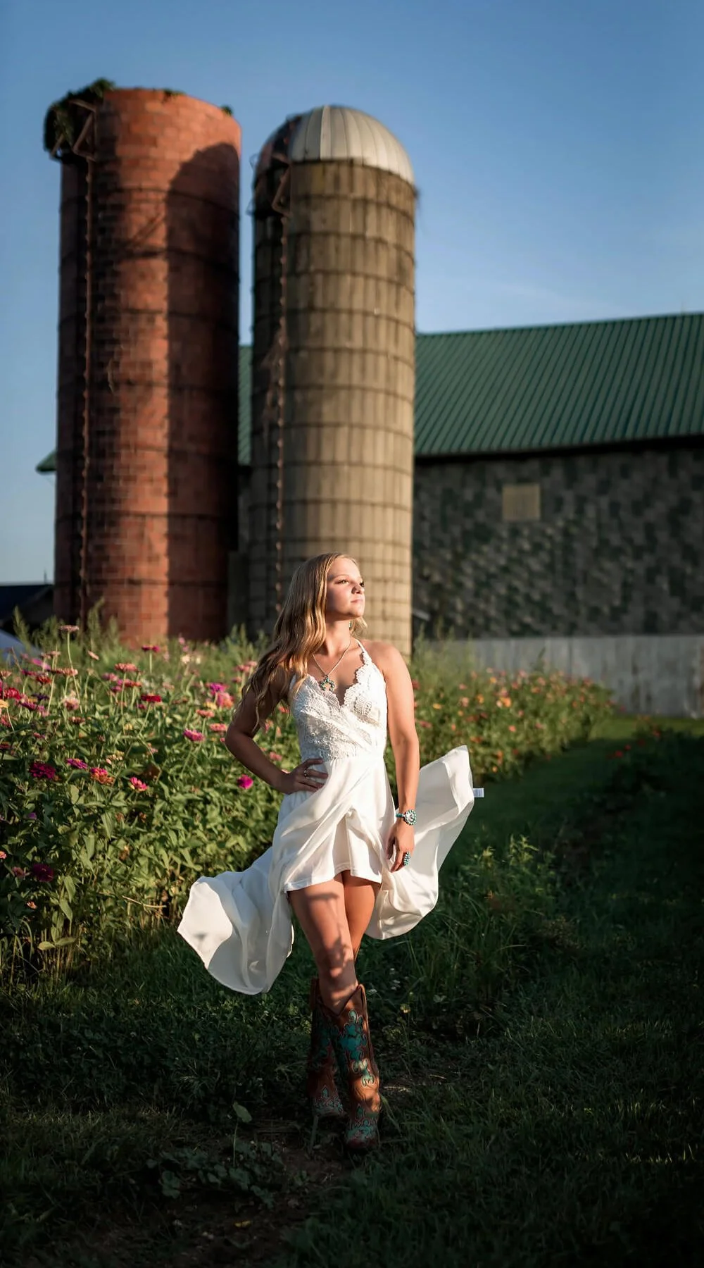 A woman in a white dress and cowboy boots standing in a field with flowers, with farm silos and a barn in the background during golden hour.