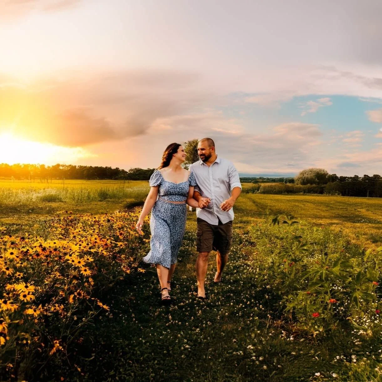 A couple walks hand-in-hand through a flower field at sunset, smiling and looking at each other.